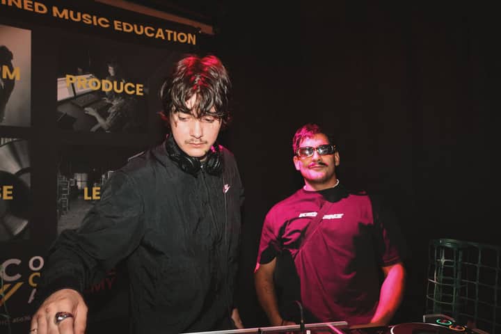 Two DJs performing at a night event with professional audio equipment and music education banner.