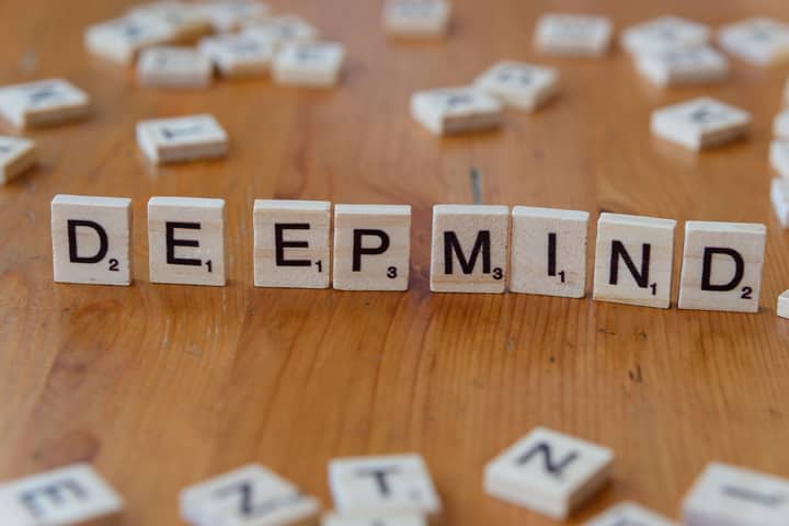 Close-up of Scrabble tiles spelling 'DeepMind' arranged on a wooden surface, with scattered tiles in the background.
