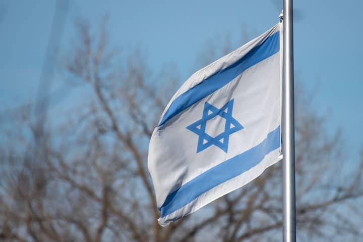 The Israeli flag featuring the Star of David against a clear blue sky and trees.