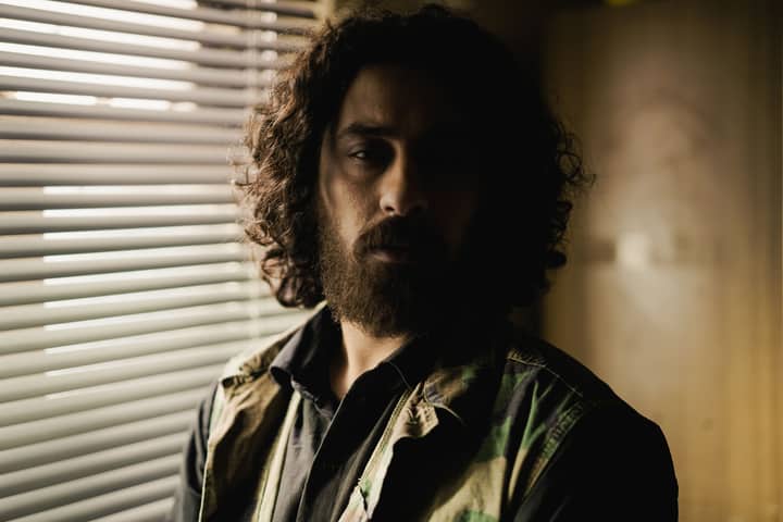 Moody portrait of a man with curly hair and beard standing by window blinds indoors.