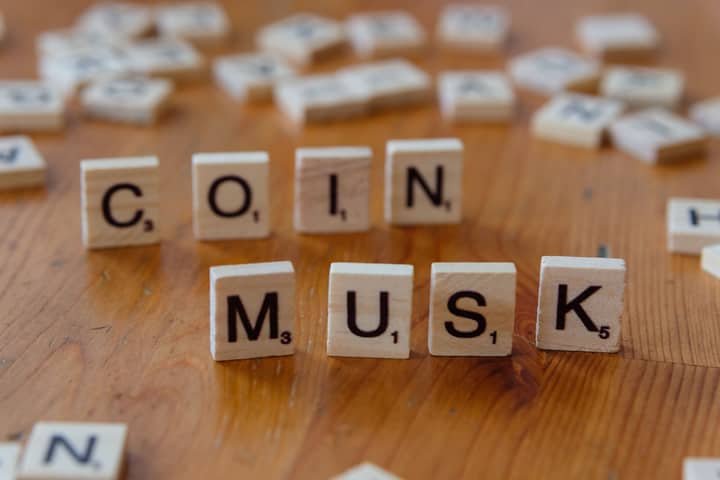 Scrabble tiles forming the words 'COIN' and 'MUSK' on a wooden table surface.