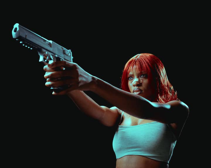 Intense portrait of a woman with red hair aiming a handgun, set against a dark background.