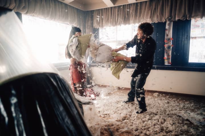 Two people enjoy a fun pillow fight indoors with feathers scattering everywhere.