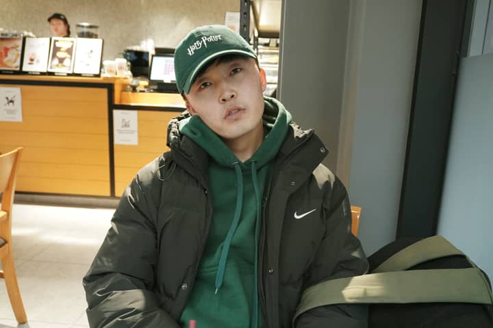 Young man with green cap and jacket sitting in a cafe, relaxed vibe.