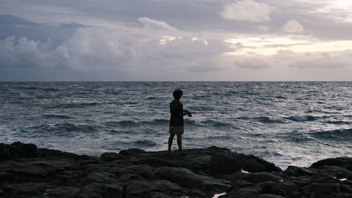 A man fishes alone on a rocky ocean shore under cloudy skies, at dusk.