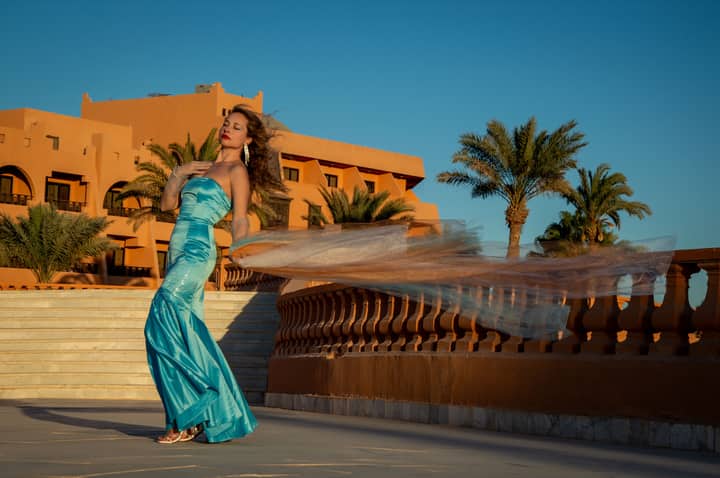 Woman in a flowing blue gown poses at a tropical resort, exuding elegance and style.