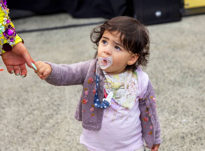 A cute toddler with a pacifier holds an adult's hand, gazing upwards in an outdoor setting.