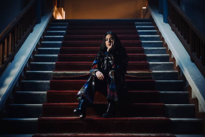 Stylish woman sitting on a red carpeted grand staircase in a moody, theatrical atmosphere.