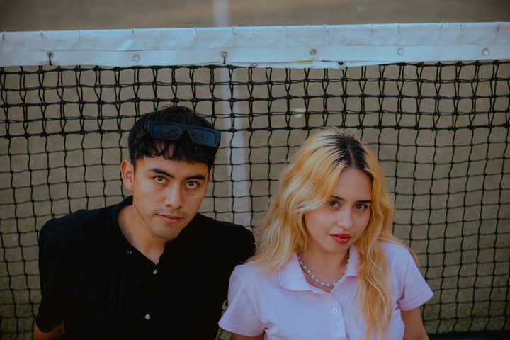 Two young adults posing by a tennis court net, showcasing casual style and fashion.
