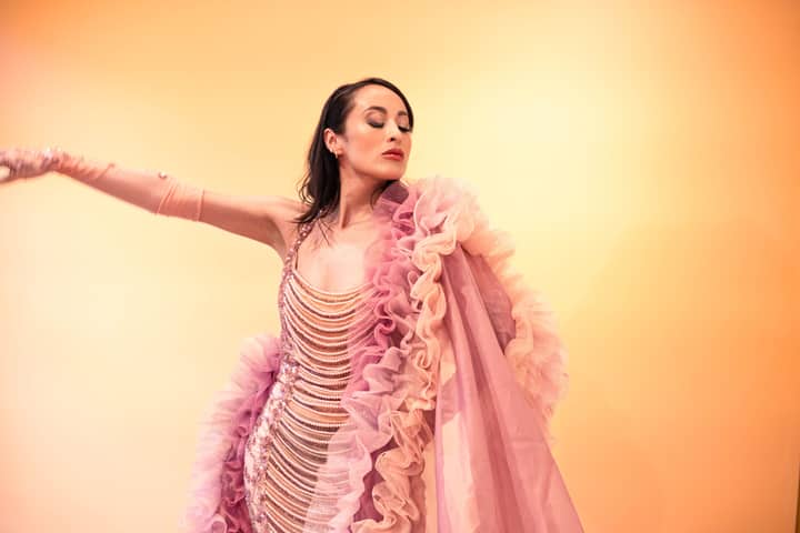 A woman gracefully poses in a ruffled pink gown against a warm pastel background.