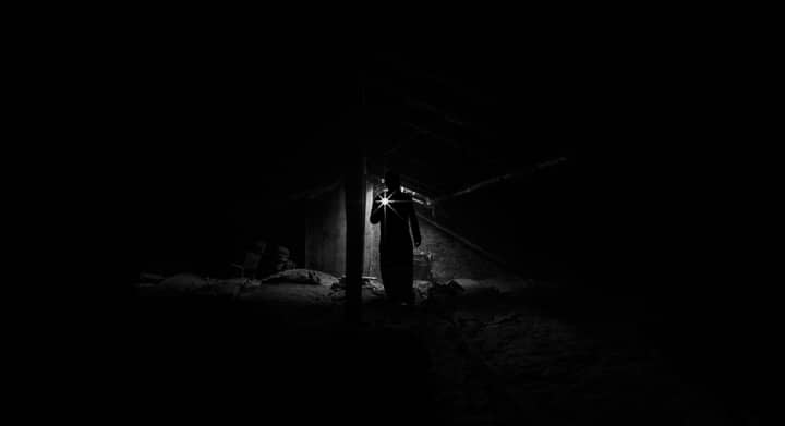 A shadowy figure explores a dimly lit, eerie attic with a flashlight.