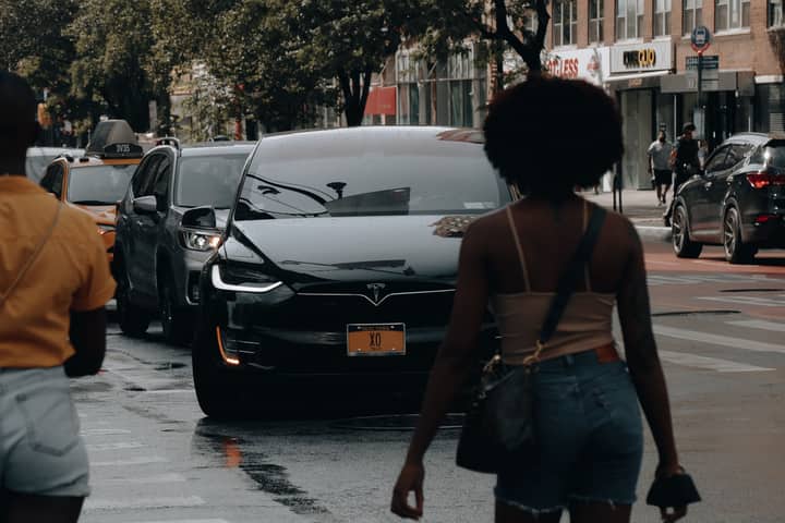 A Tesla car navigates through traffic on a busy street in New York City with pedestrians nearby.
