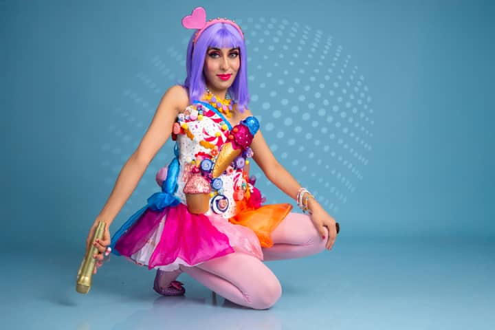 Woman in a vibrant candy-themed costume with purple wig and heart accessory.