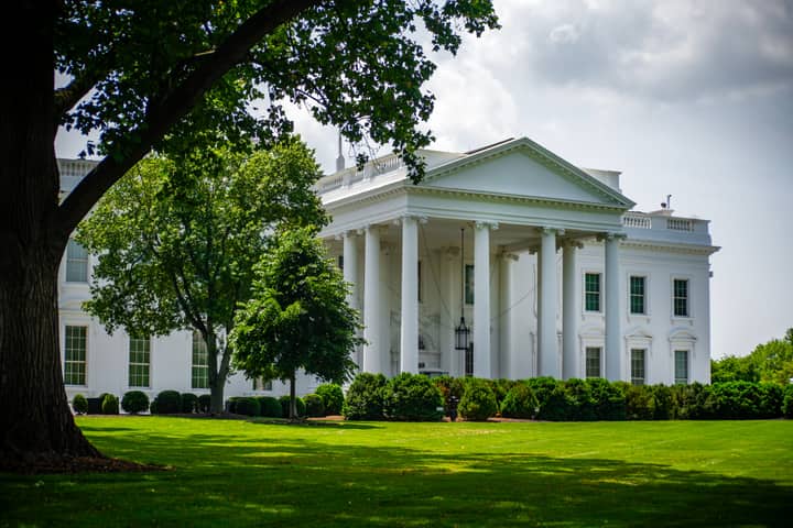 The White House framed by trees and greenery, in Washington, D.C., under a bright sky.