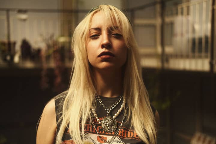 Blonde woman with rock style necklace and graphic tee in outdoor setting.