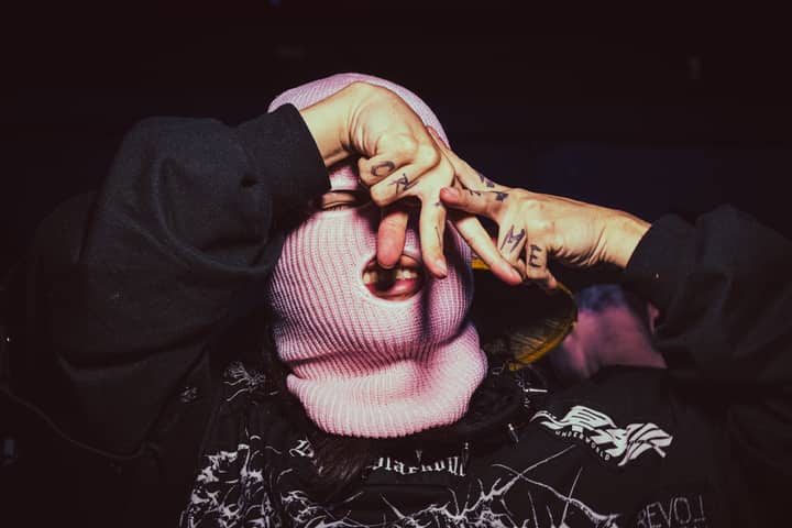A person wearing a pink ski mask with tattoos on fingers in a dark, mysterious setting.