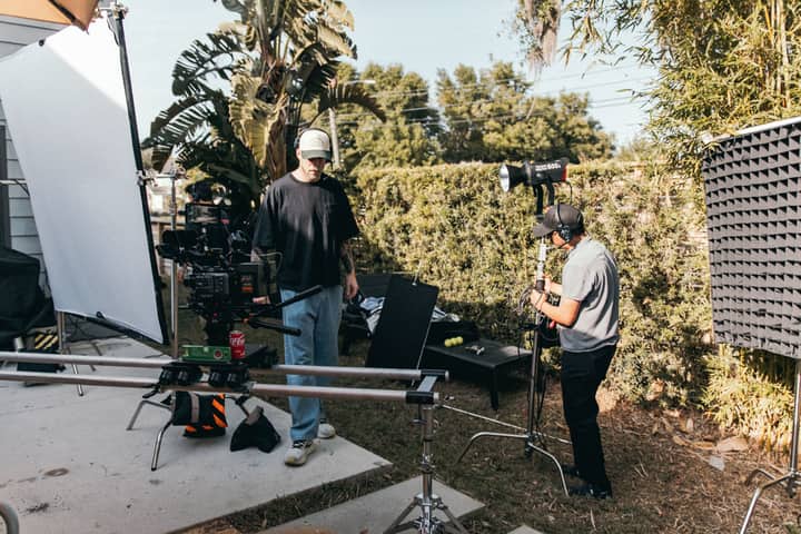 A film crew setting up lights and camera equipment outdoors with lush greenery.