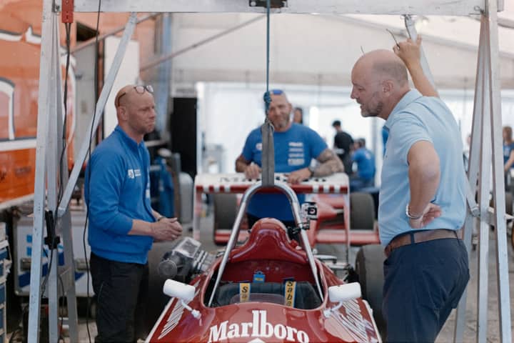 Team members discuss strategy beside a classic race car in a garage setting.