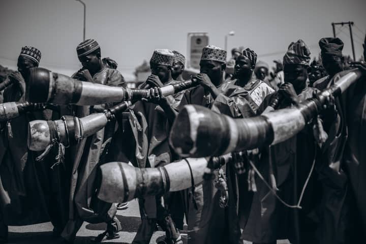 Black and white photo of African musicians playing traditional horns during a vibrant street parade.