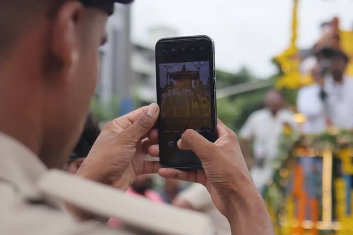 A person photographs the colorful Jagannath Rath Yatra parade with a smartphone.