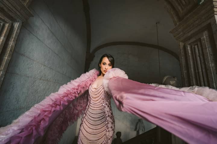 Stylish woman in a dramatic pink gown stands elegantly inside a historic building.