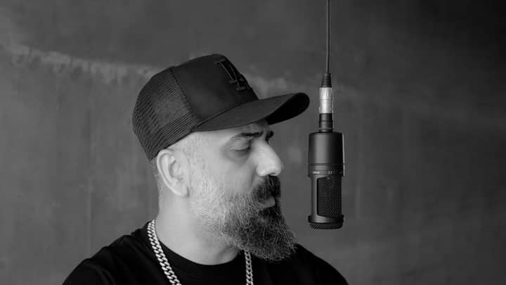 Black and white portrait of a bearded rapper wearing a cap in front of a microphone.