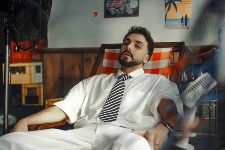 A stylish man in a white suit and tie relaxes indoors with vintage decor elements.