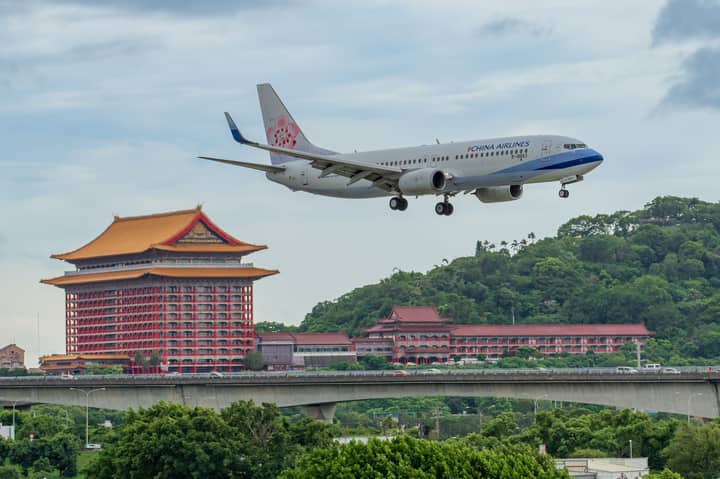China Airlines plane approaches landing over Grand Hotel Taipei amidst lush greenery and cloudy sky.