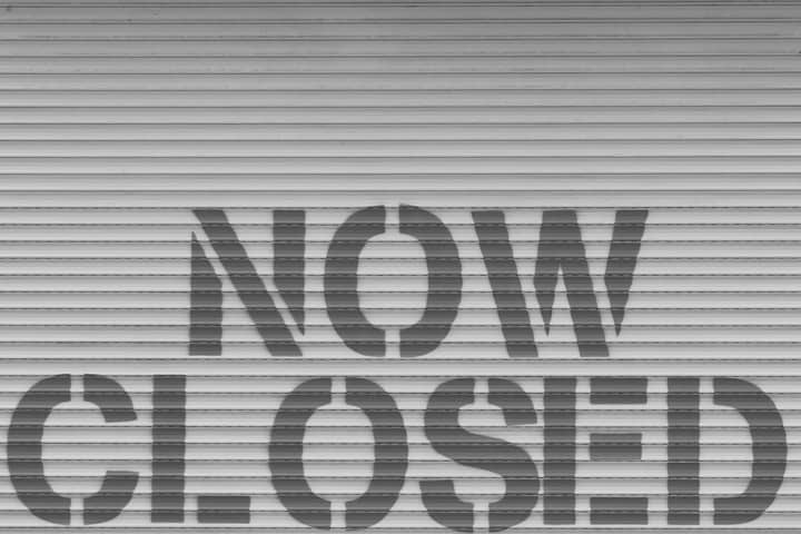 Black and white image of a 'Now Closed' sign on a steel shutter, symbolizing closure.