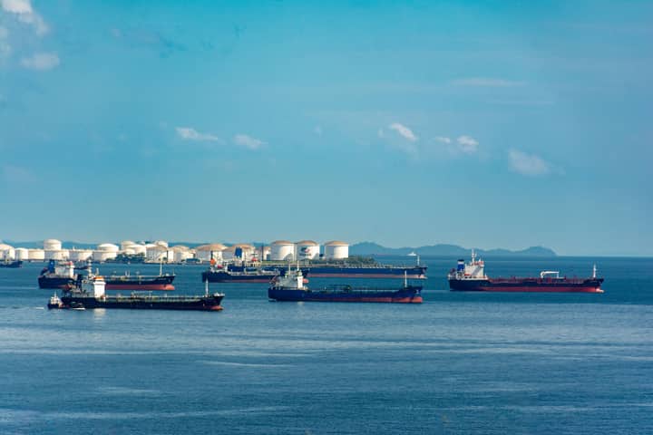 A fleet of cargo ships docked near oil storage tanks along a serene coastline with a clear blue sky above.