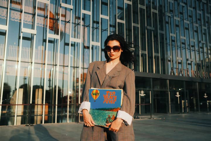 Fashionable woman with sunglasses and vinyl album standing outside a modern glass building at sunset.