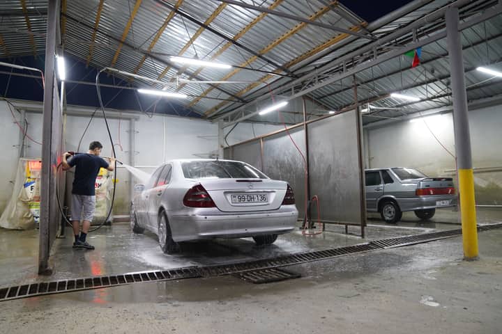 A man uses a pressure washer to clean a silver sedan in a covered car wash at night.
