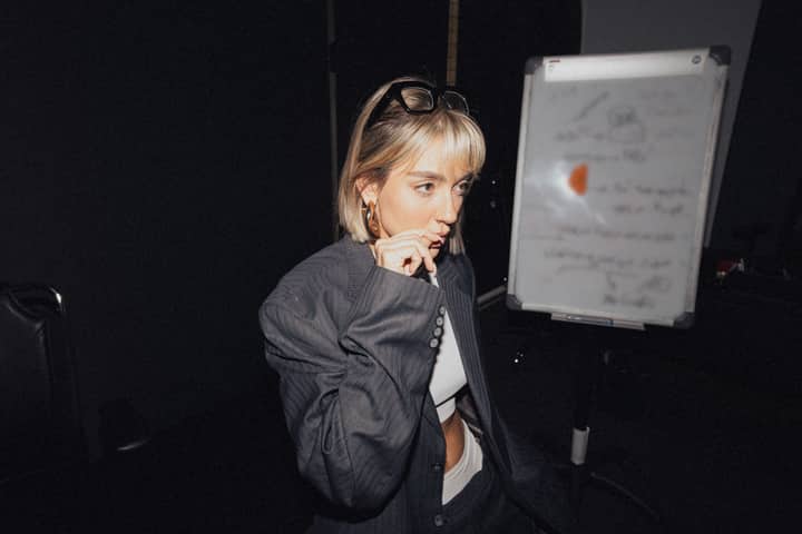 Young woman thoughtfully standing in a dimly lit office near a whiteboard.