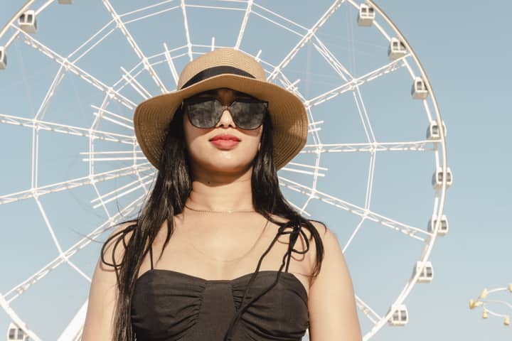 Stylish woman in hat and sunglasses poses near Ferris wheel in Baku on a sunny day.