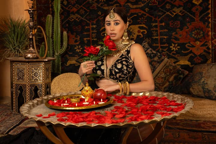 A woman in traditional attire holding roses, surrounded by lavish decor and jewelry.