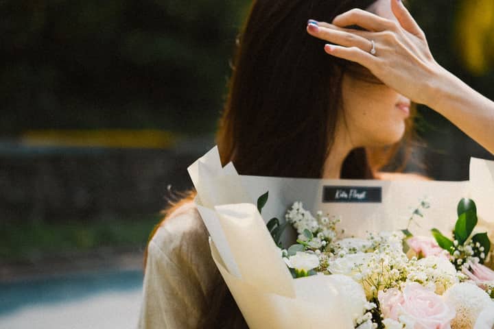 A woman with long hair holds a bouquet of soft-colored flowers outdoors, creating an elegant and serene atmosphere.