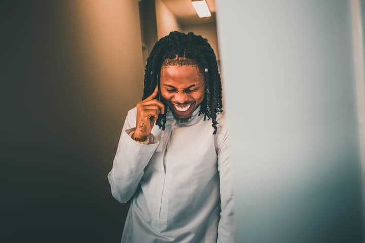 A happy man with braided hair smiling while standing indoors, wearing a fashionable white jacket.