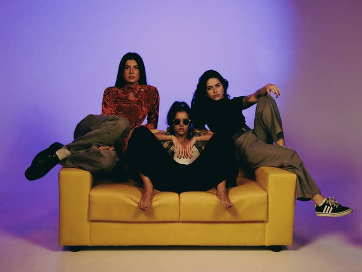 Three stylish women pose confidently on a yellow couch against a vivid background.