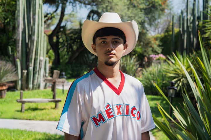 A young man wearing a cowboy hat and México jersey stands amidst lush greenery.