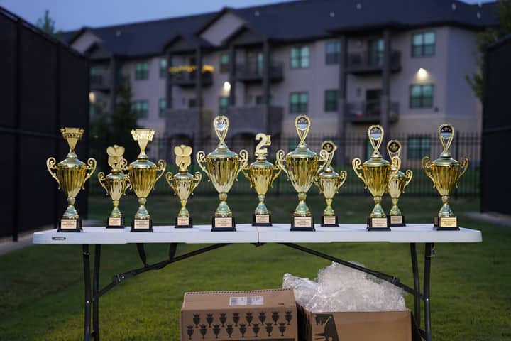 Lineup of gold trophies on a table outdoors with apartment backdrop.
