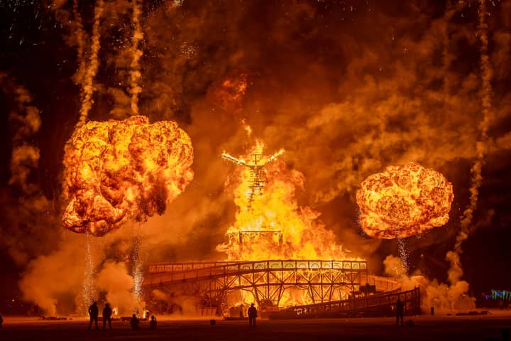 Dramatic fire and fireworks display at Burning Man festival with glowing effigy silhouette.