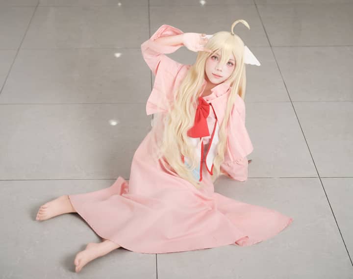 Cosplay character sitting indoors in a pink dress with a playful pose and long blonde hair.