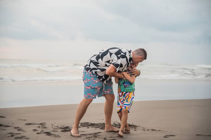A joyful interaction between father and son playing on a sandy beach under a cloudy sky.