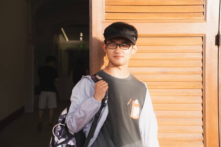 A young man with glasses stands in sunlight, leaning against a wooden door, exuding a casual and relaxed vibe.