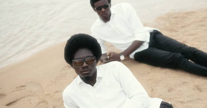 Two stylish men in sunglasses relax on a sandy beach in minimalistic attire.