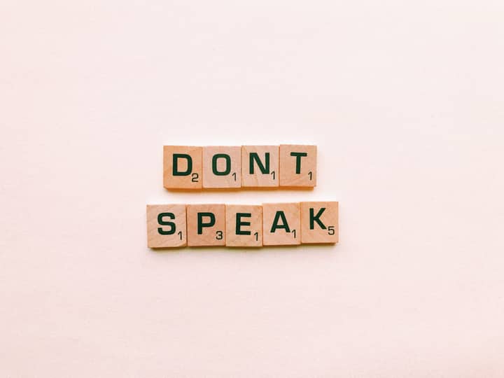 Wooden Scrabble tiles spelling 'Don't Speak' on a pink background, emphasizing silence.