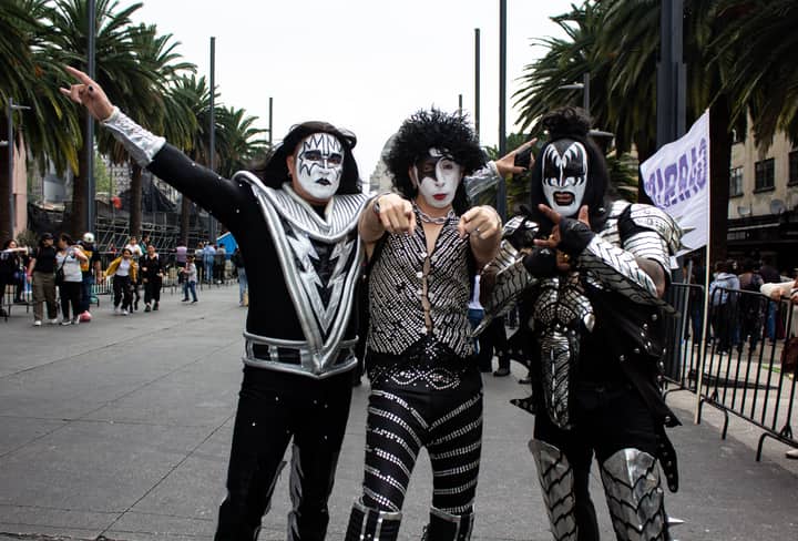Three adults dressed as KISS band members pose in a lively street scene in Mexico City.
