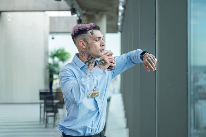 Young tattooed man in stylish shirt enjoys a drink on a city balcony at dusk.