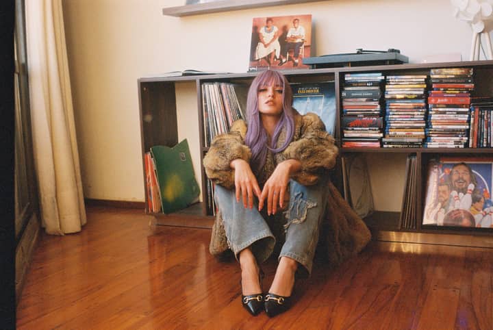 Stylish woman with lavender hair sitting among records and CDs in a cozy room.