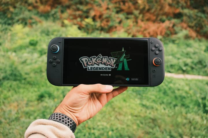 A hand holding a handheld gaming console displaying the Pokémon Legends game screen outdoors.